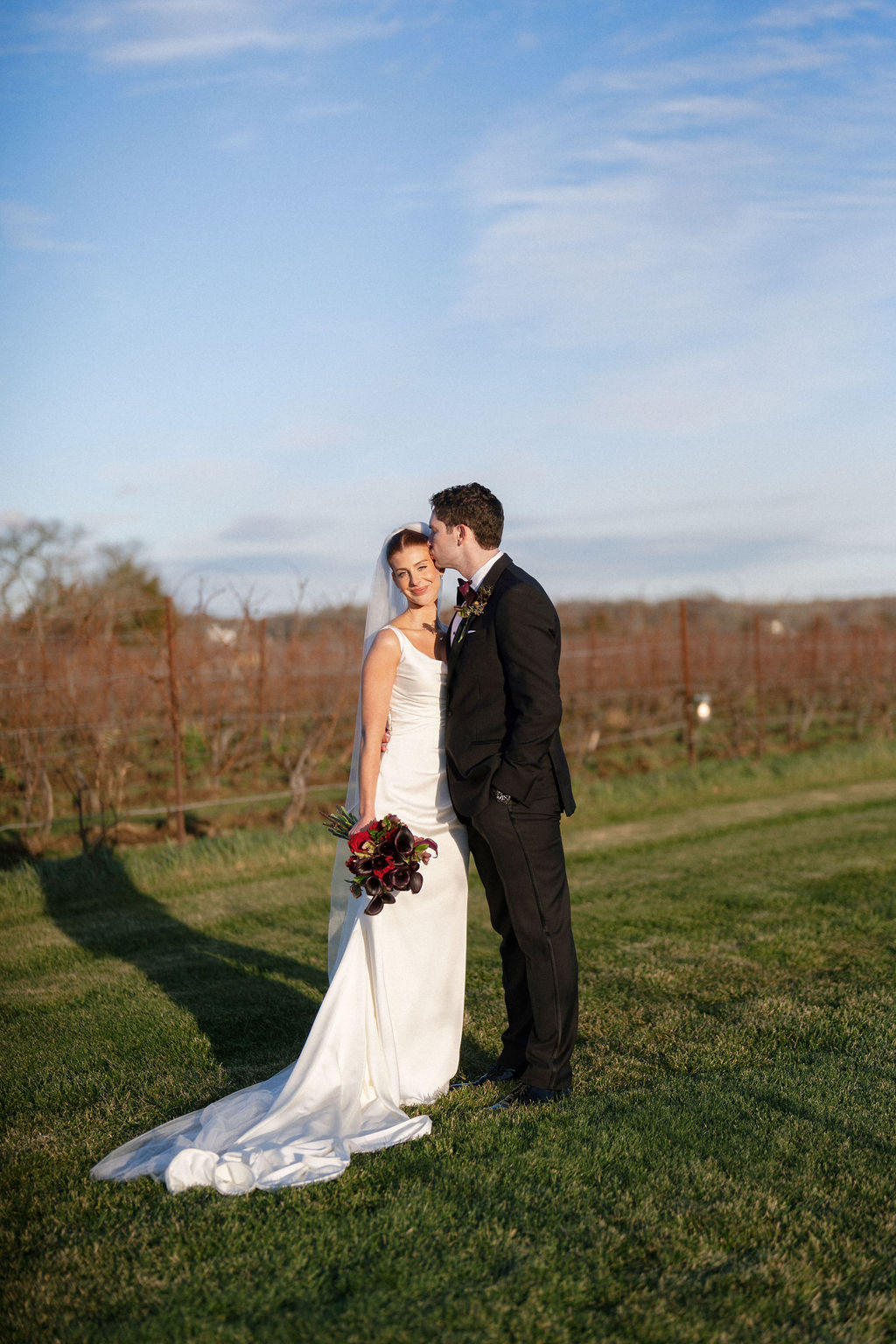 Bride and groom at Saltwater Farm Vineyard in Stonington, Connecticut holding a moody winter wedding bouquet in burgundy and plum tones with evergreens and calla lilies.