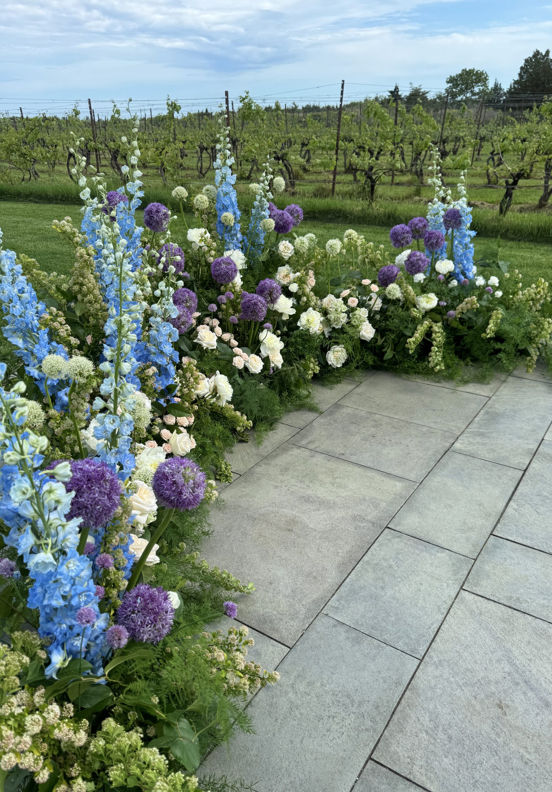 Ceremony flowers at Saltwater Farm Vineyard featuring purple allium, blue delphinium, ivory roses, and lush greenery in a vineyard setting.